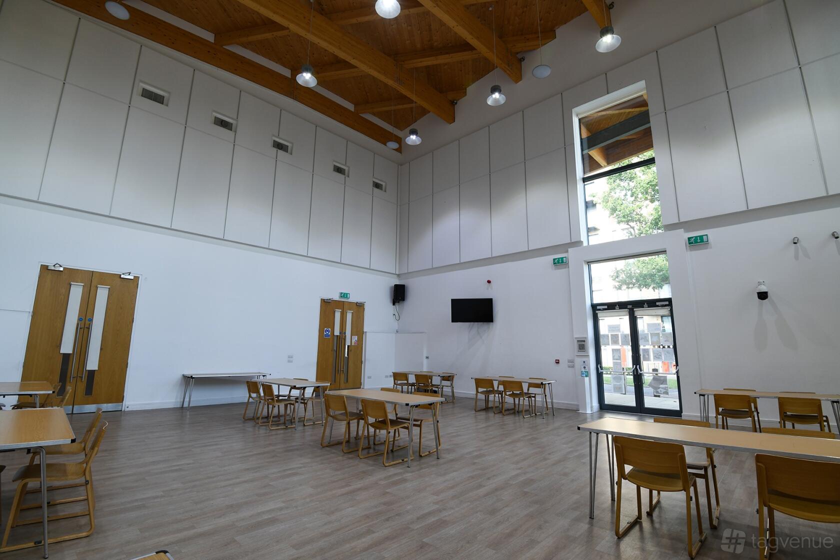 A community hall with high wooden beam ceilings, large windows, and scattered tables at OneStonegrove Wilberforce Hall.